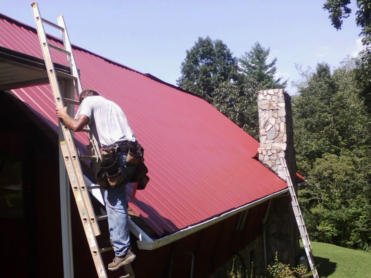 Roofer on a ladder beside a red metal roof and stone chimney — Soffit & Fascia Repair by Roofing Brothers in Batavia